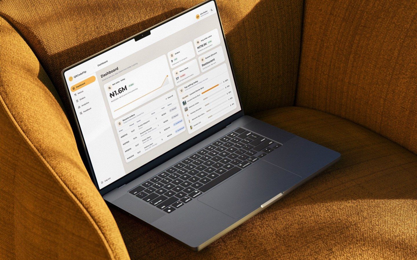 The QRCodeFlip admin dashboard on a laptop resting on a mustard armchair: today’s net sales, orders, menu views, the busiest QR point, recent orders and top sellers