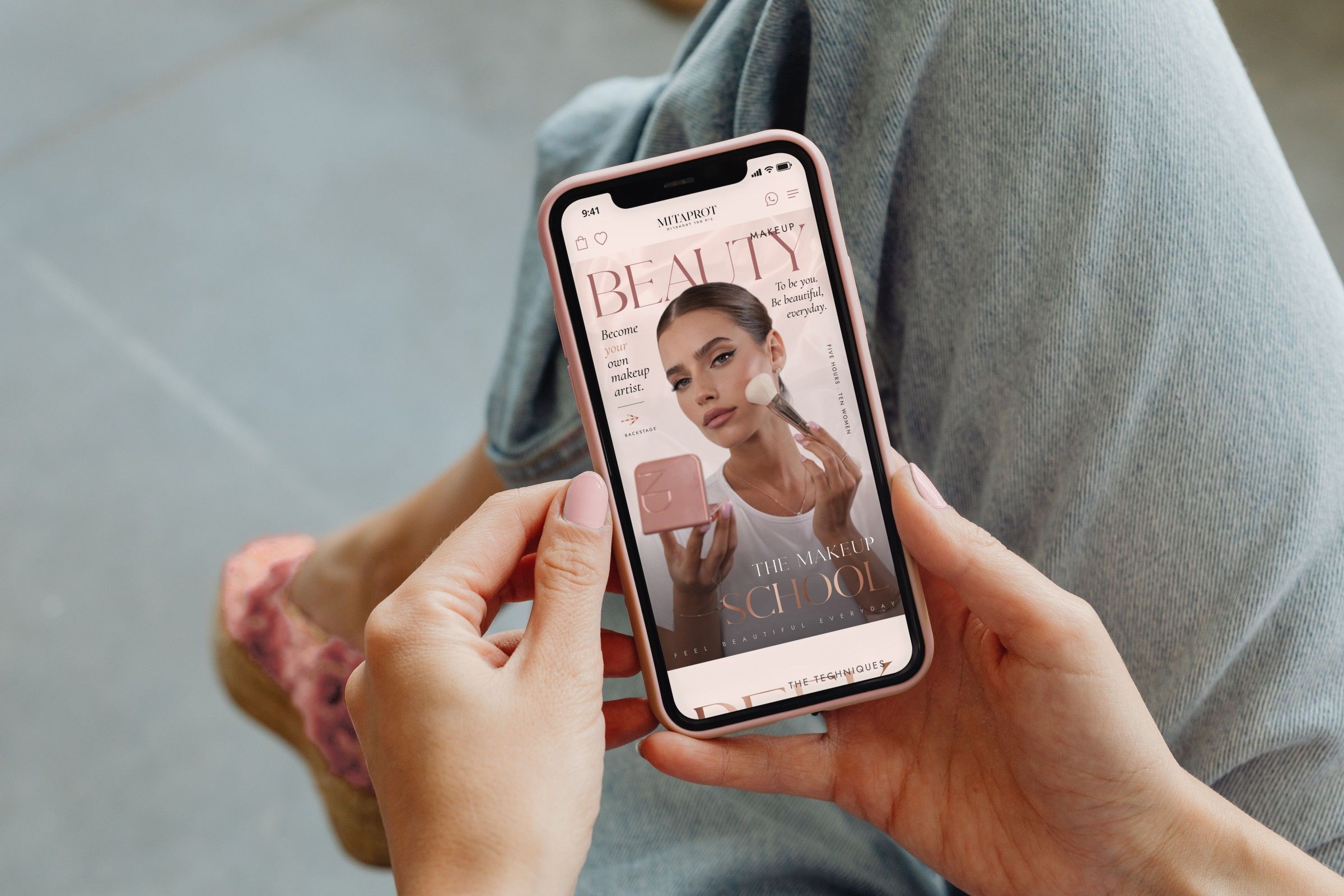 The home page hero on an iPhone in a blush pink case, held in two hands with pale pink nails over denim: Beauty in large letters, “Become your own makeup artist”, a model with a brush and a pink compact, and The Makeup School