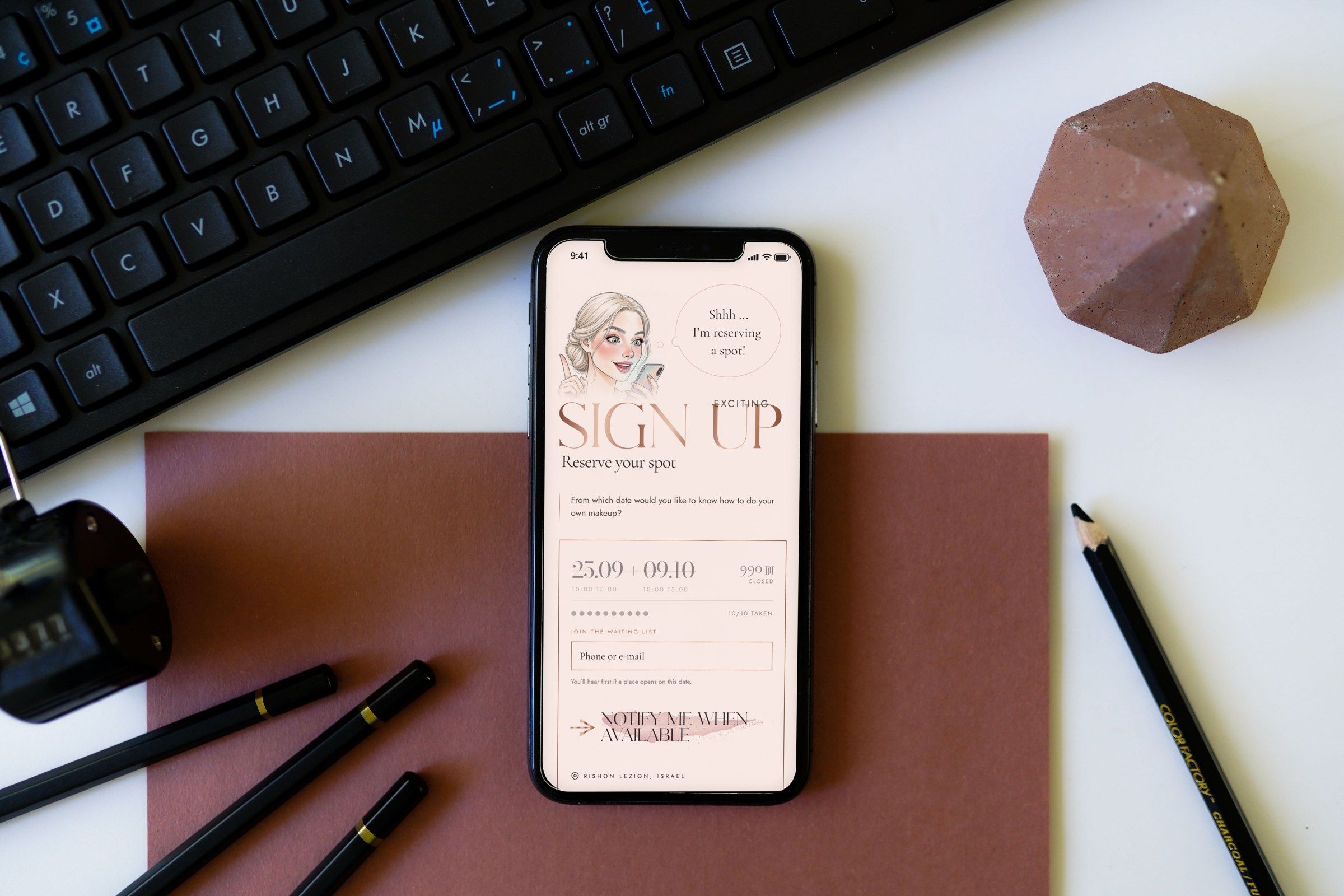 The sign-up part of the home page on an iPhone lying on a desk beside a keyboard and pencils: the illustrated girl saying “Shhh… I’m reserving a spot!”, the Sign up heading and a full course date with its waiting-list field