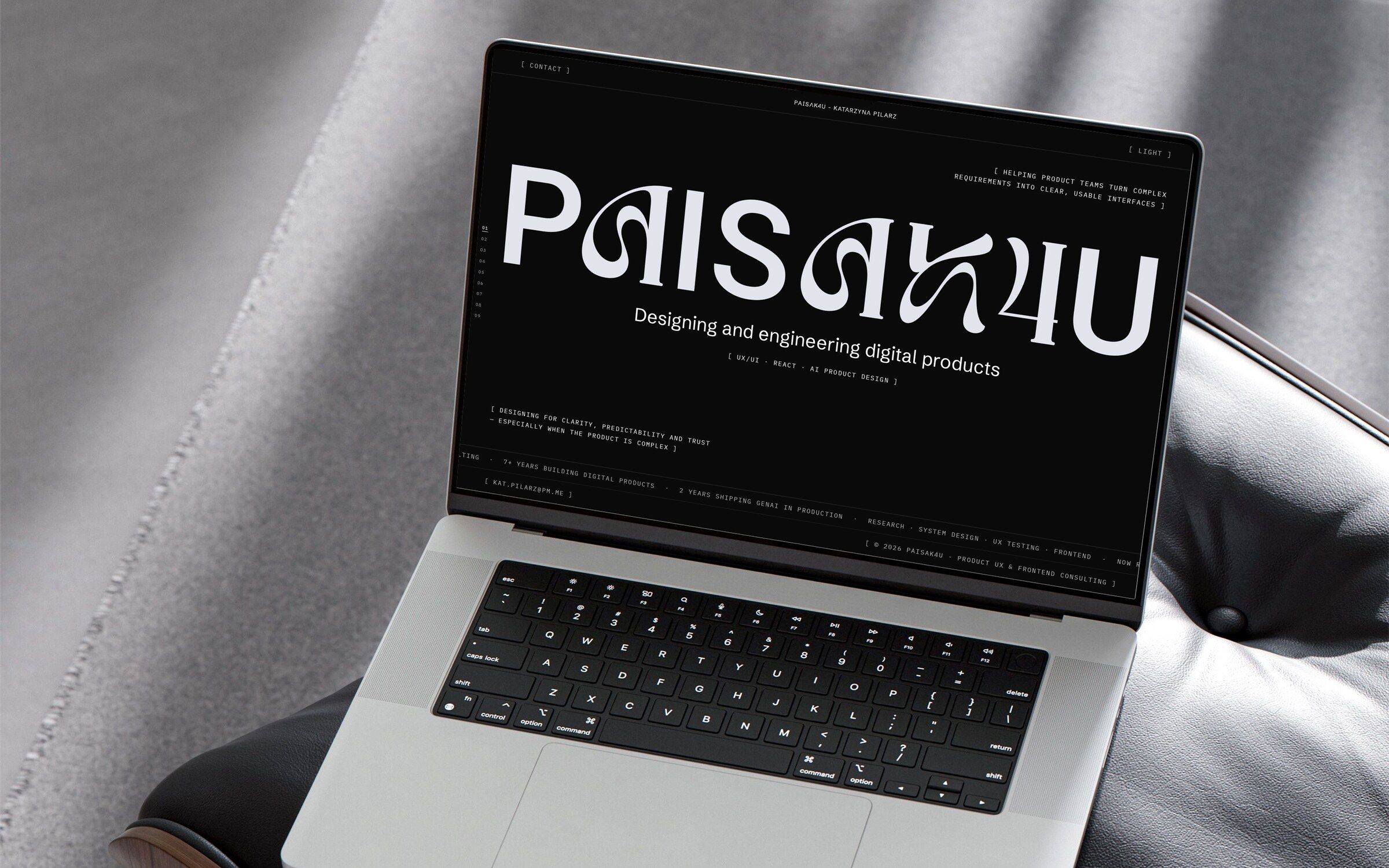 The PAISAK4U home page in the dark theme on a laptop resting on a black leather lounge chair