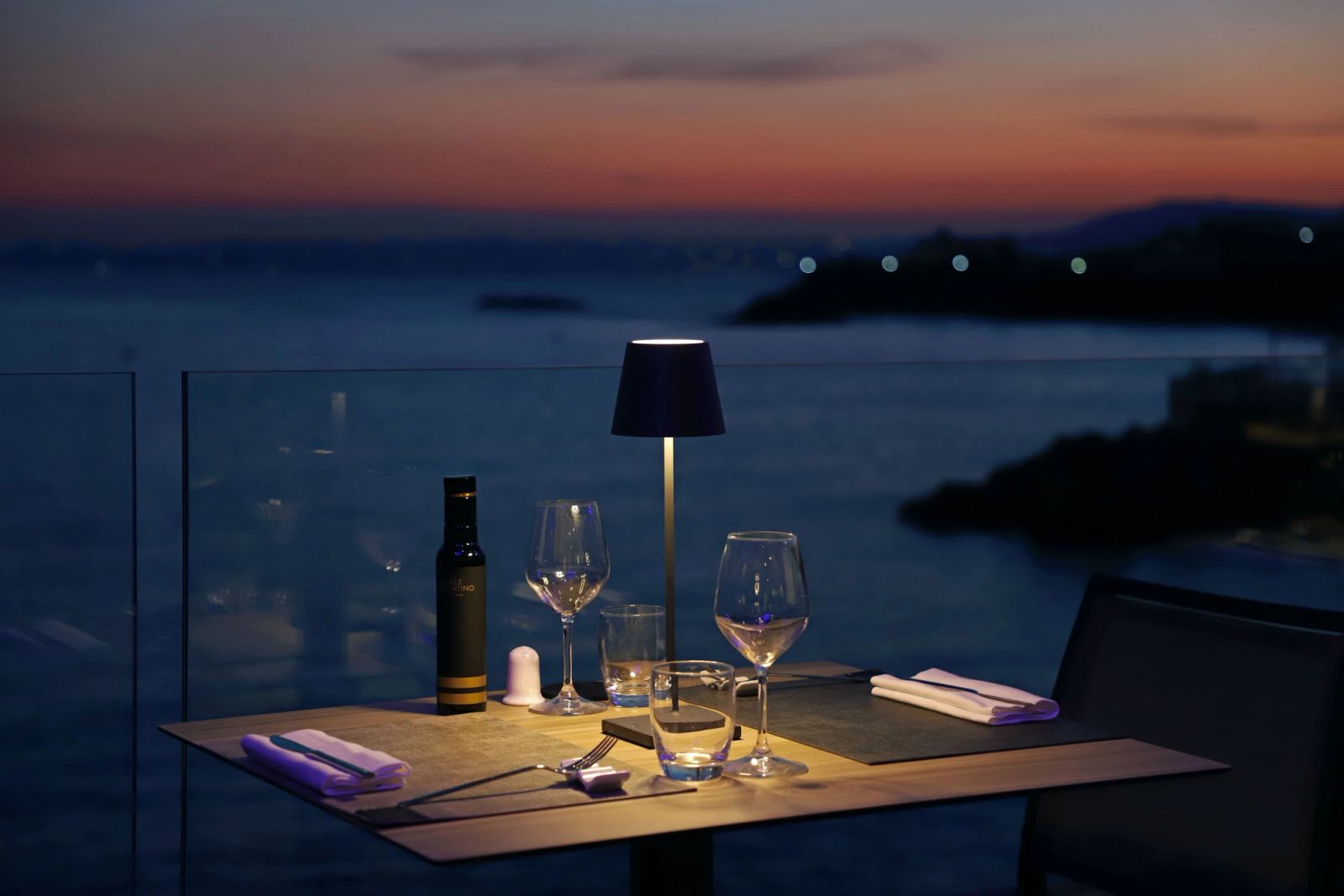 Tavolo apparecchiato per due su una terrazza sul mare al tramonto, con lampada accesa e calici