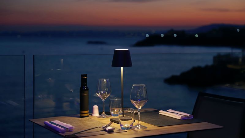 Tavolo apparecchiato per due su una terrazza sul mare al tramonto, con lampada accesa e calici