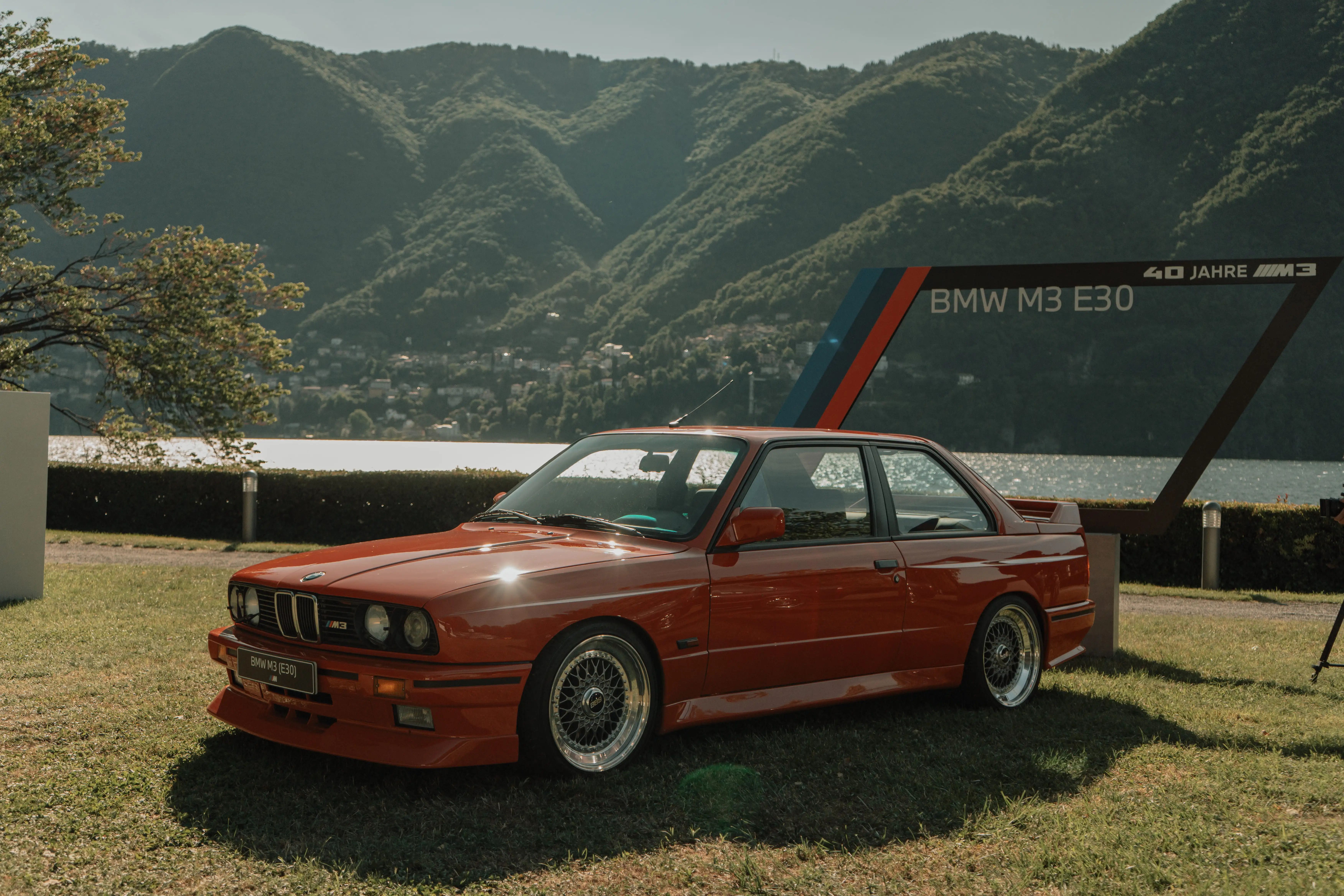 BMW M3 E30 rossa sul prato davanti al lago e alle montagne, con la cornice «40 Jahre M3»