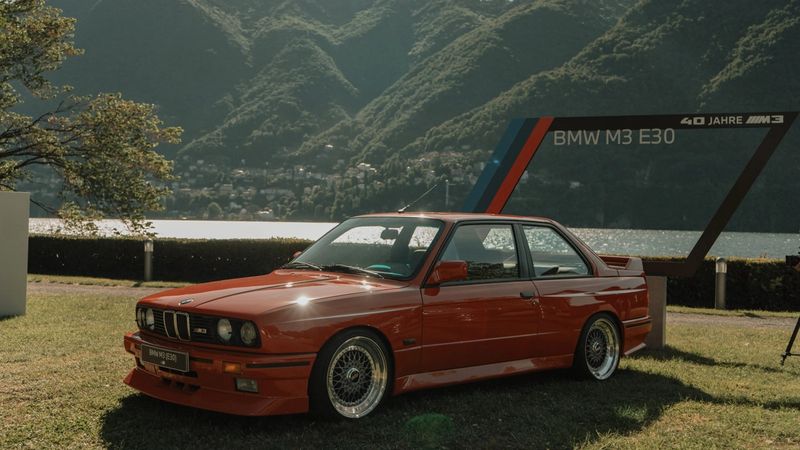 BMW M3 E30 rossa sul prato davanti al lago e alle montagne, con la cornice «40 Jahre M3»