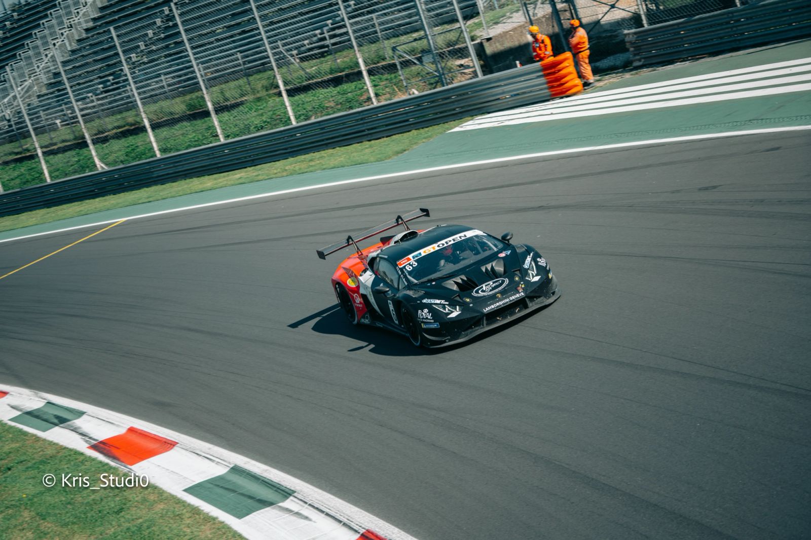 Auto GT3 nera e arancione in curva sul cordolo tricolore, con due commissari di pista in tuta arancione