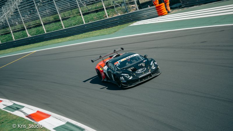 Auto GT3 nera e arancione in curva sul cordolo tricolore, con due commissari di pista in tuta arancione