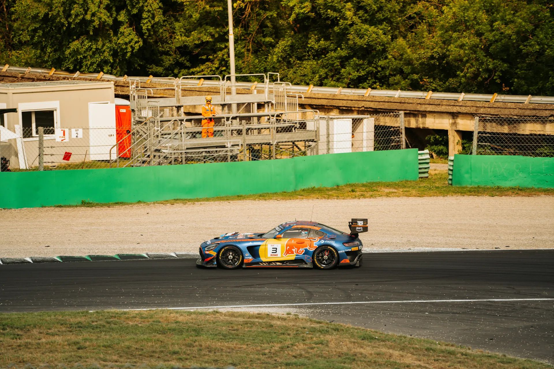 Auto GT3 blu e arancione con il numero 3 in pista, dietro un muretto verde e un commissario in tuta arancione