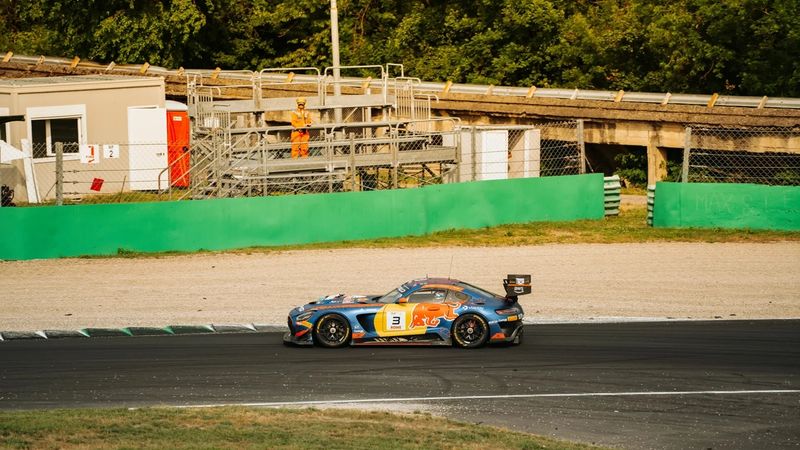 Auto GT3 blu e arancione con il numero 3 in pista, dietro un muretto verde e un commissario in tuta arancione
