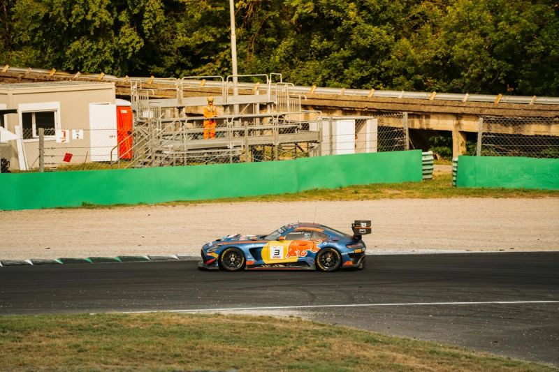 Auto GT3 blu e arancione con il numero 3 in pista a Monza, dietro un muretto verde e un commissario in tuta arancione