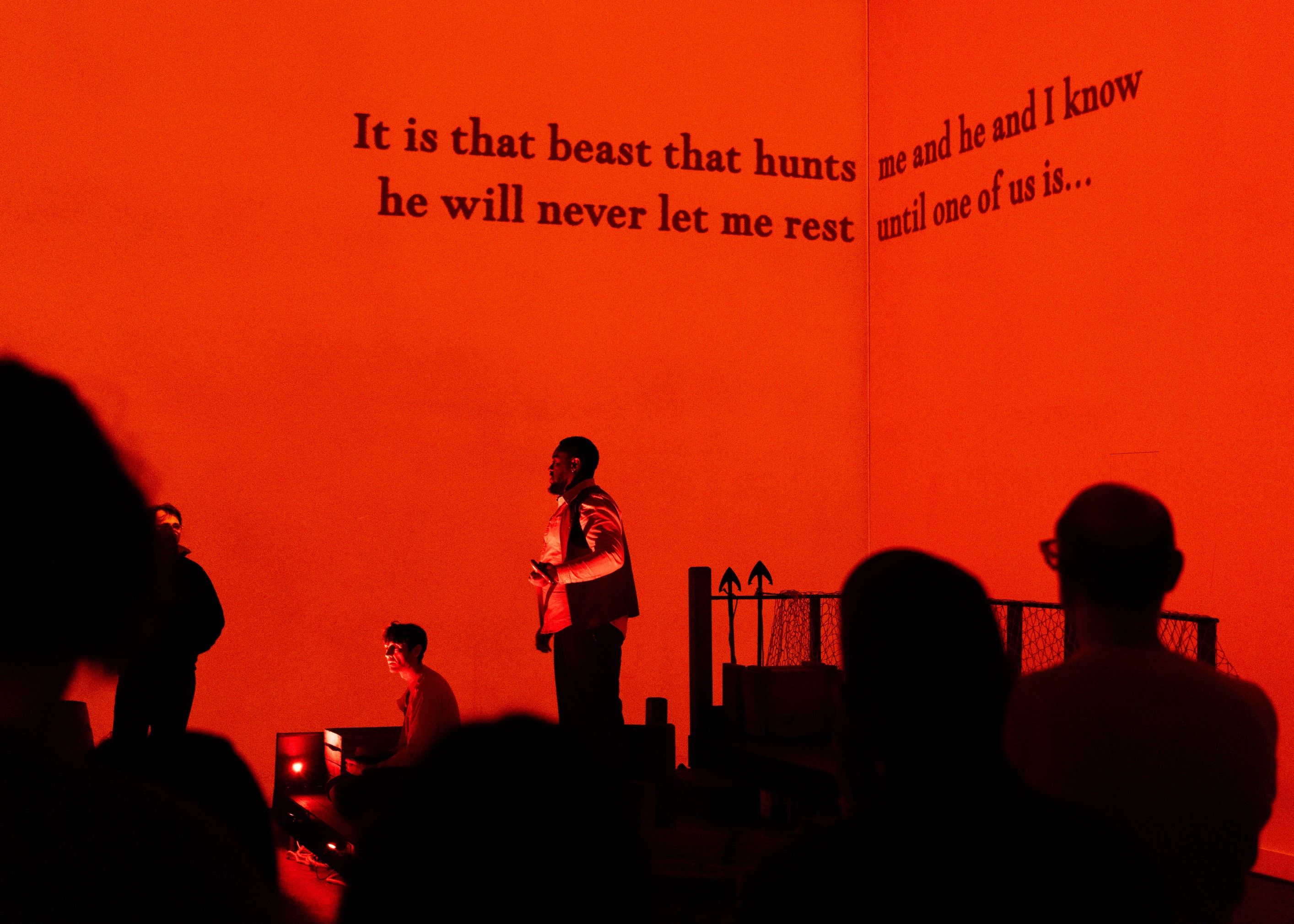 Man on stage with red backdrop and text written on screen behind him - accessible creative captions for deaf, deafened and hard of hearing audiences