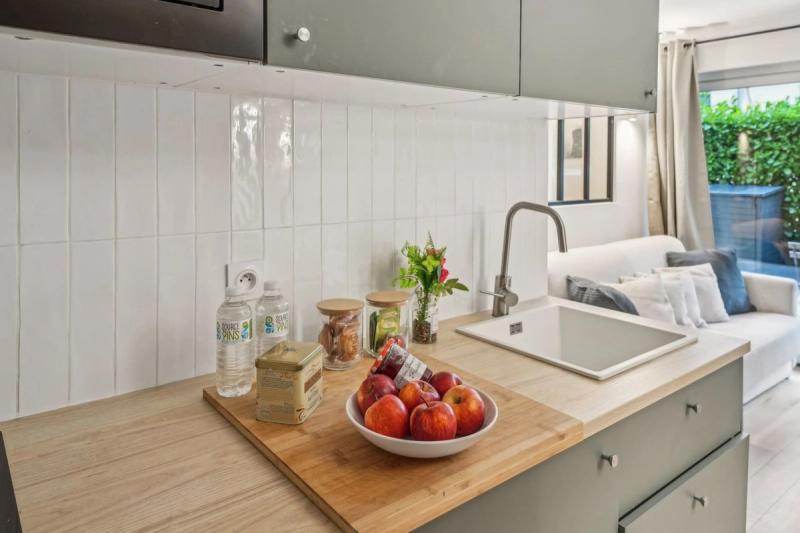 Kitchen counter with a bowl of apples, white tiled backsplash, and sage green cabinets facing a sofa near garden doors.