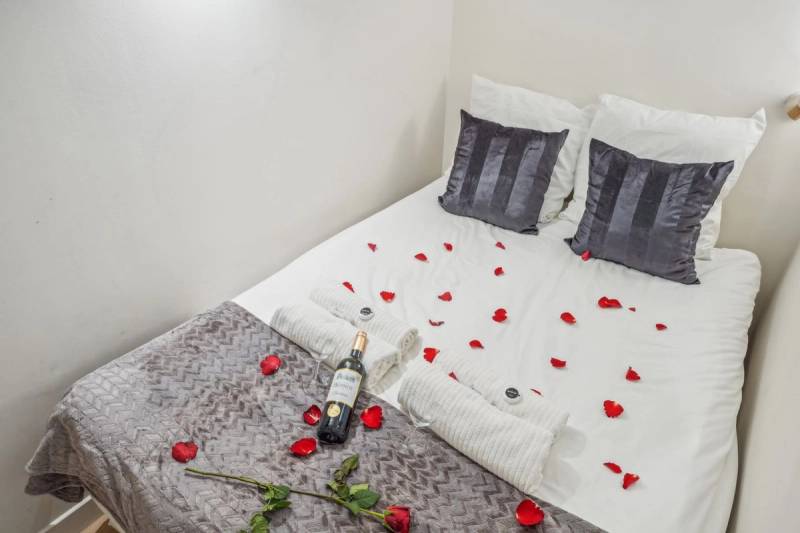 Double bed scattered with red rose petals, a wine bottle, and rolled white towels.