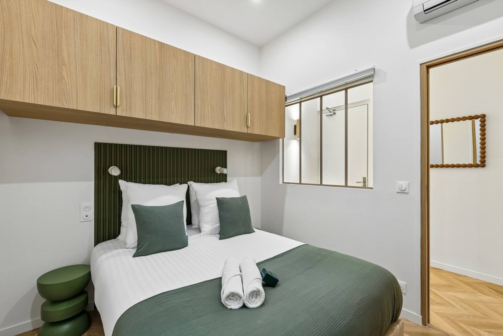 Bedroom with a fluted green headboard, overhead oak cabinets, and an interior glazed window.