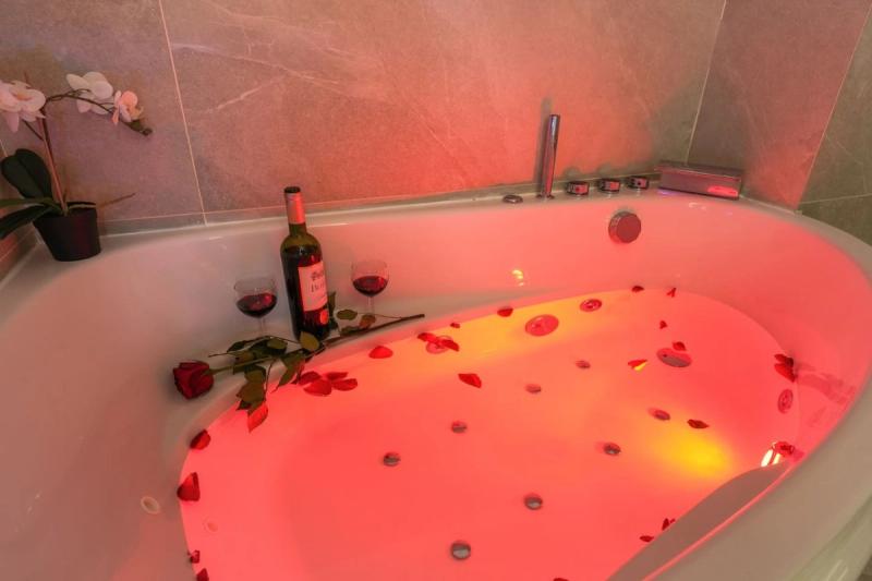 Jacuzzi tub lit red with scattered rose petals, a wine bottle, two glasses, and an orchid on grey stone tiles.