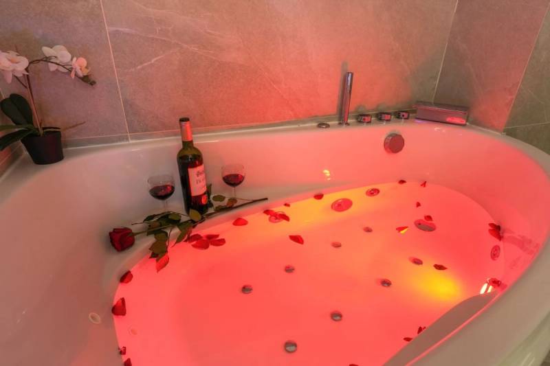 Jacuzzi tub lit red with scattered rose petals, a wine bottle, two glasses, and an orchid on grey stone tiles.
