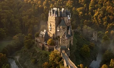 Eltz Castle rises on a rock in the forest, surrounded by the Elzbach stream, its distinctive towers lit in golden light