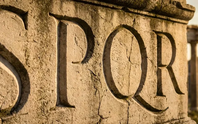 Die Buchstaben SPQR sind in einen verwitterten antiken römischen Steinbogen eingemeißelt, im Hintergrund das Forum Romanum