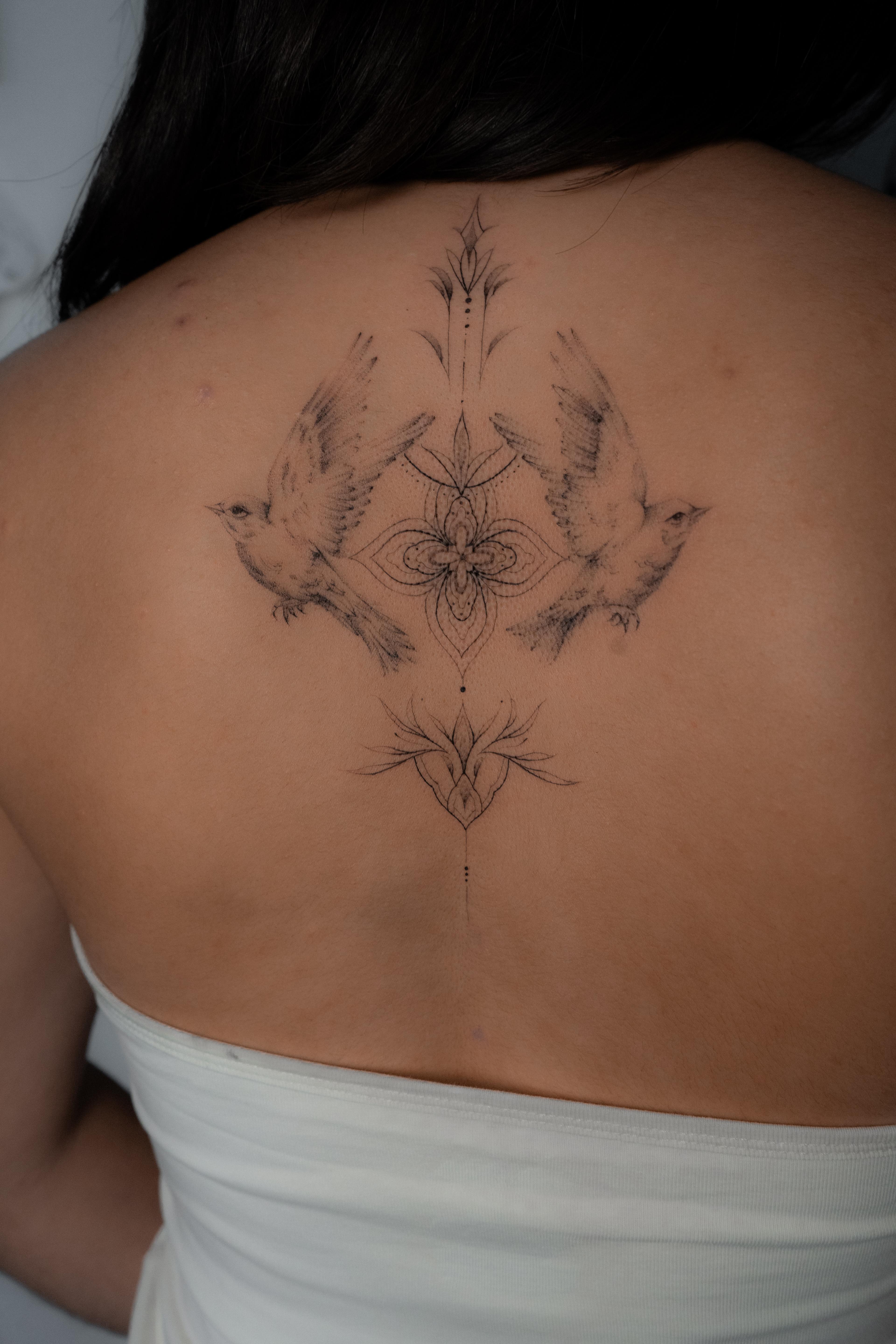 Two fine-line birds framing a symmetrical ornamental upper-back tattoo
