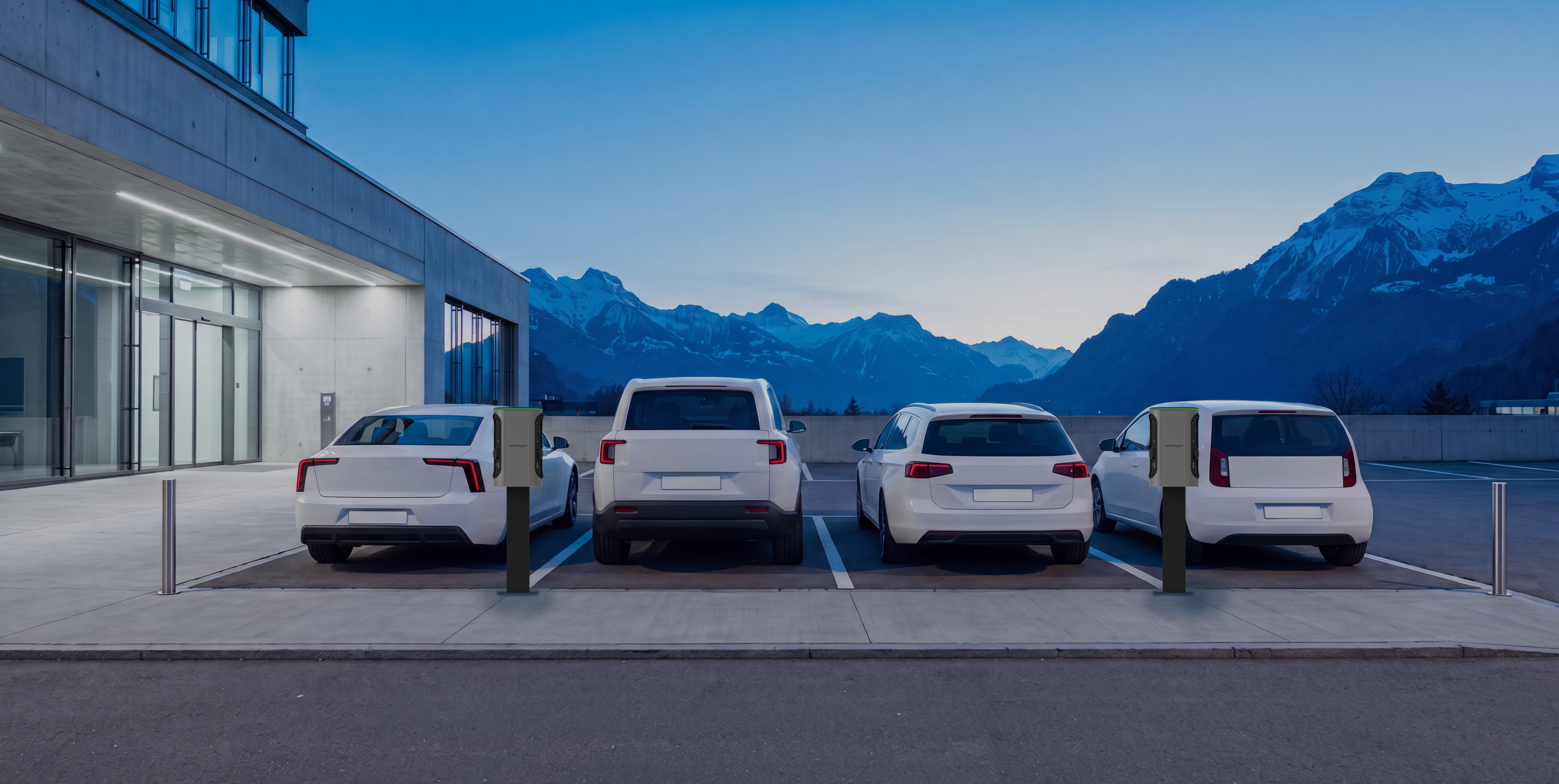 Firmenparkplatz in der Dämmerung: vier weisse Fahrzeuge an Ladesäulen vor einem Bürogebäude, dahinter Berge