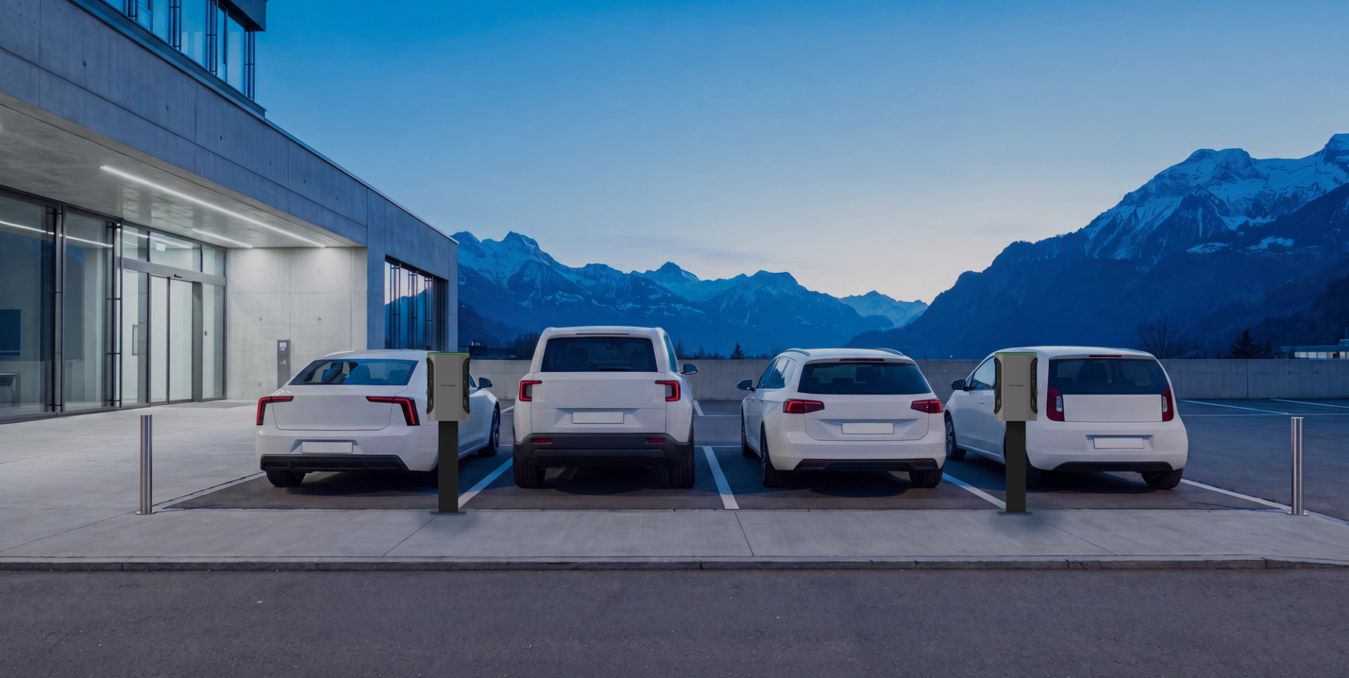 Firmenparkplatz in der Dämmerung: vier weisse Fahrzeuge an Ladesäulen vor einem Bürogebäude, dahinter Berge