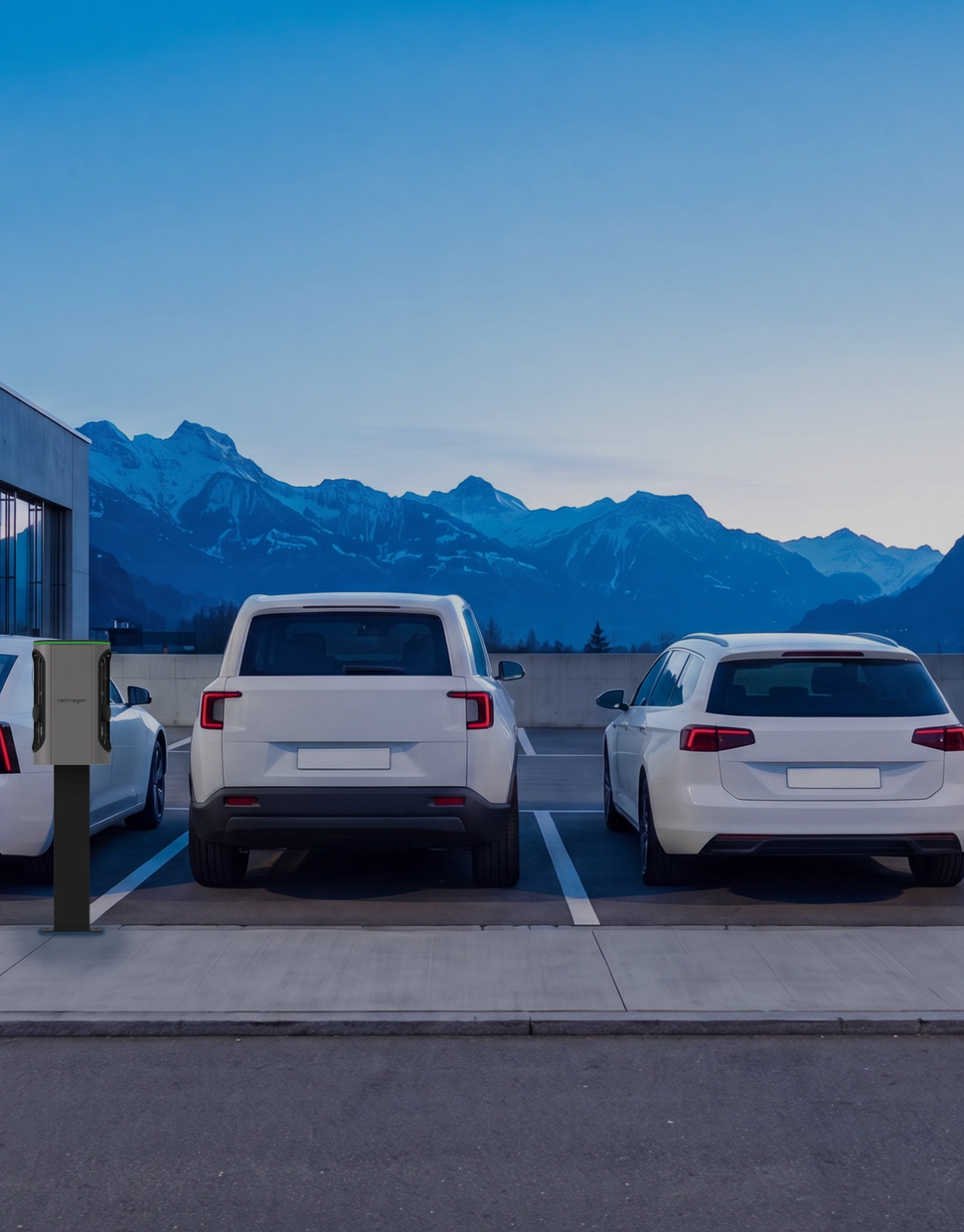 Firmenparkplatz in der Dämmerung: vier weisse Fahrzeuge an Ladesäulen vor einem Bürogebäude, dahinter Berge