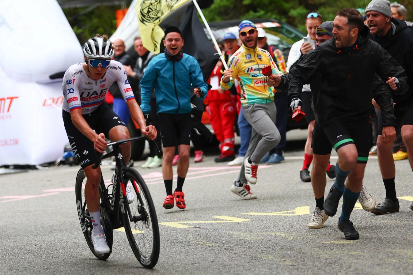 In the foreground, Tadej Pogačar rides to stage victory. In the background, a jubilant fan pays his own homage to Marco Pantani, sunglasses and all
