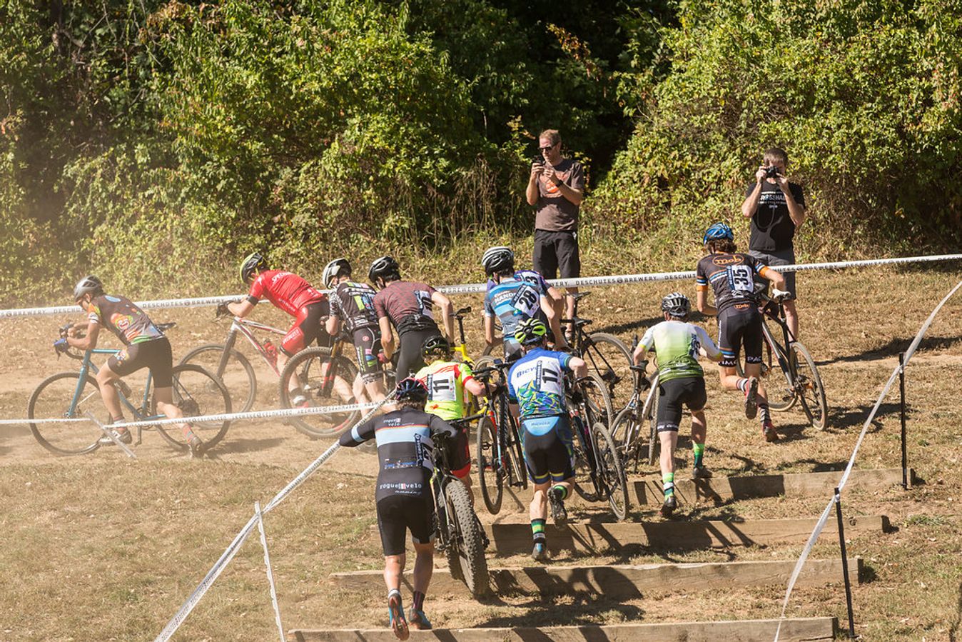 Riders clamber up the steps on a dusty cyclocross section