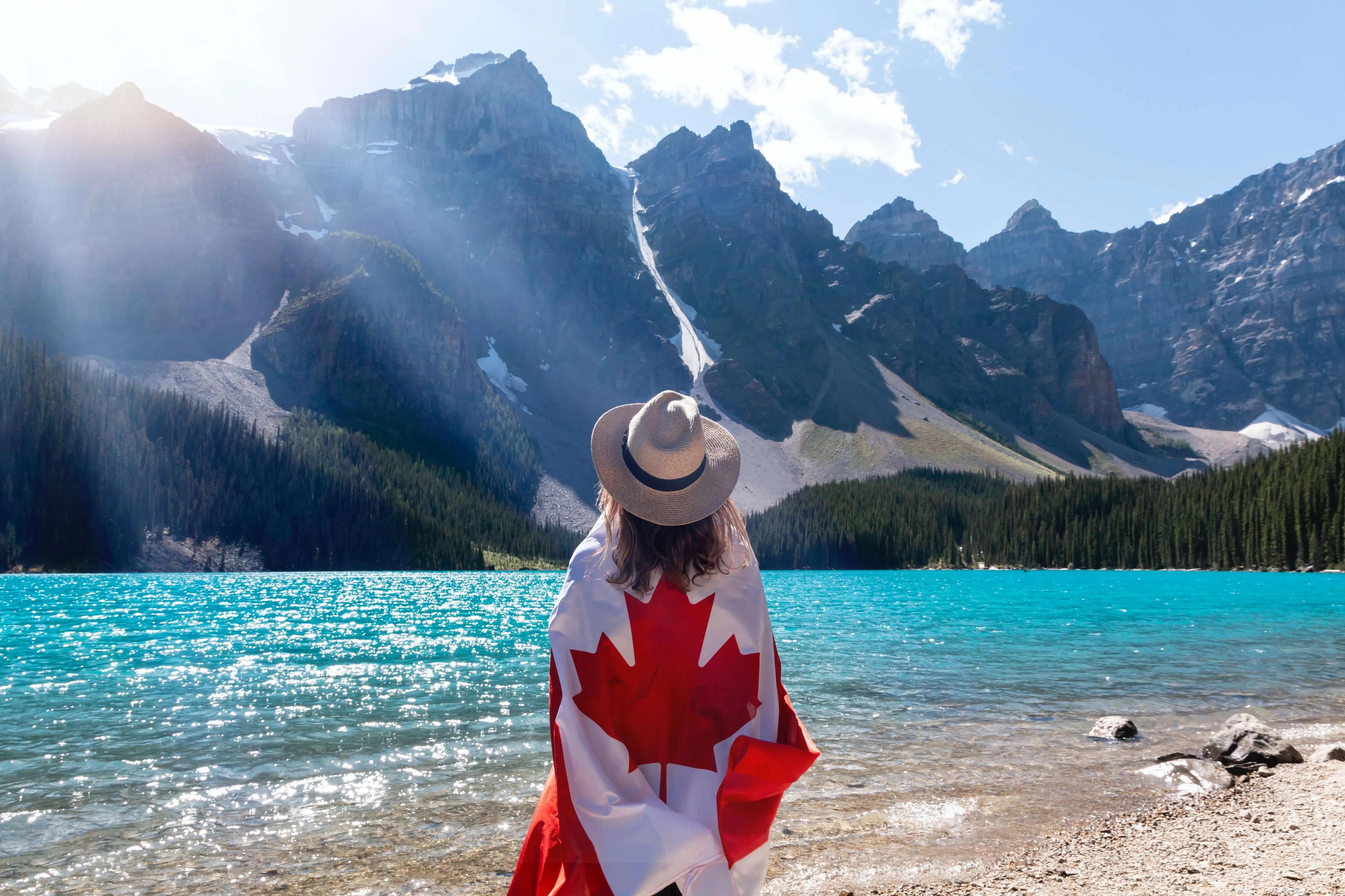 A person in a brimmed hat draped in a Canadian flag looking across a blue lake at a mountain