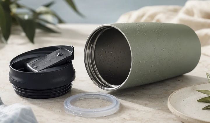 A travel mug separated into its body, lid and seal on a clean tabletop.