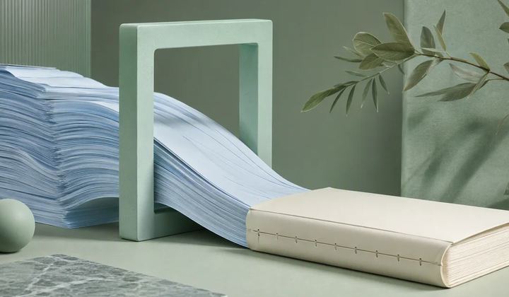 A stream of pale blue paper passes through a mint frame toward a bound ivory book.