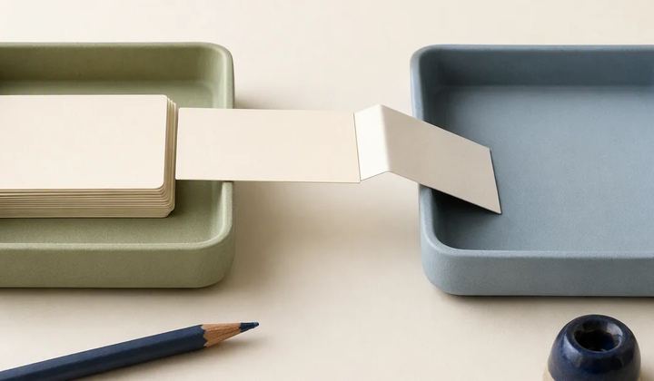 A folded paper strip connects a stack of blank cards in a green tray to an empty blue tray.