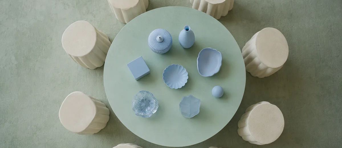 An overhead composition of ivory stools surrounds a mint circular table holding a small set of distinct pale blue objects.