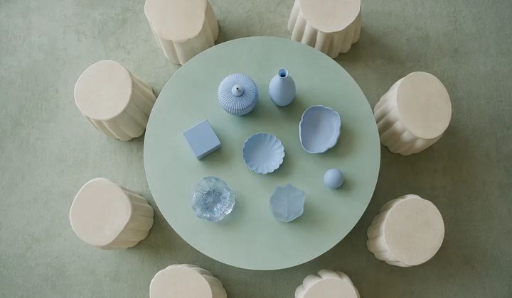 An overhead composition of ivory stools surrounds a mint circular table holding a small set of distinct pale blue objects.