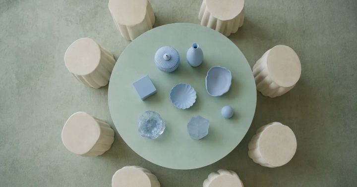 An overhead composition of ivory stools surrounds a mint circular table holding a small set of distinct pale blue objects.