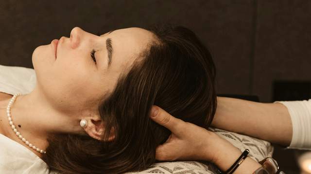 Woman lying on a patterned pillow with her eyes closed while a therapist massages the back of her head