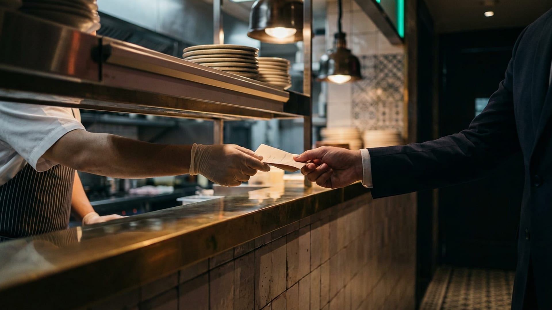 Chef passing a folded note across a brass kitchen pass to a maitre d' in a dark suit