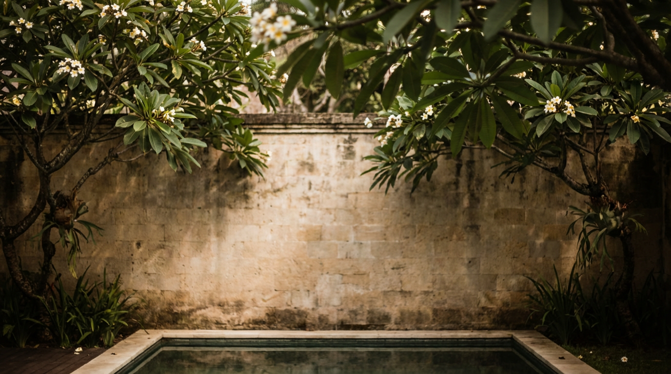 High garden wall enclosing a villa pool with mature frangipani trees and deep shadow on the stone perimeter
