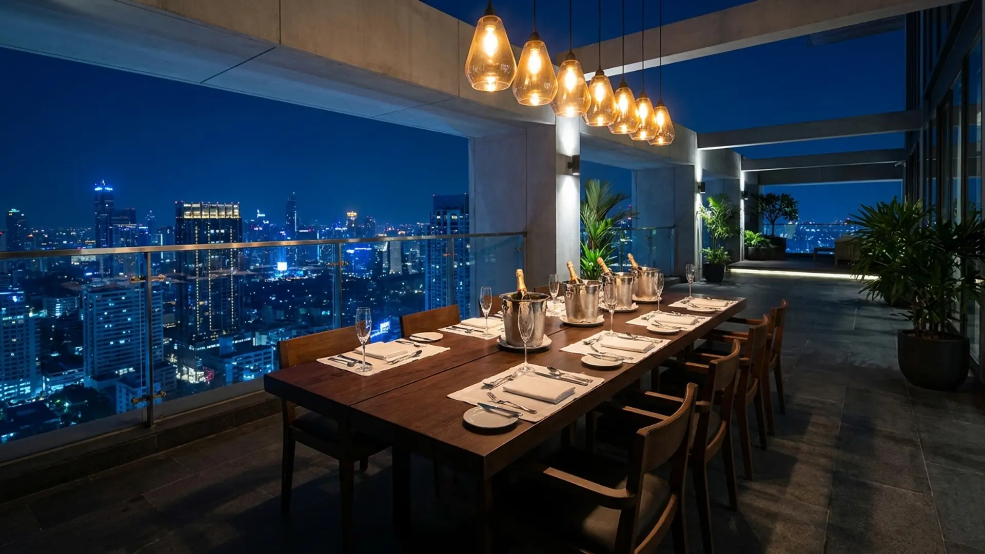 A long teak dinner table on a private terrace set for eight with low brass candleholders and the Bangkok skyline in the background