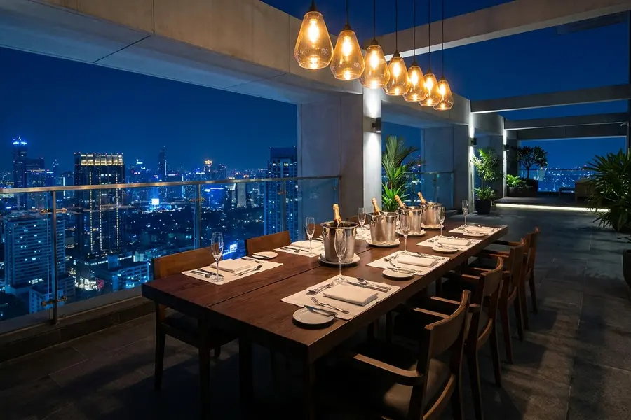 A long teak dinner table on a private terrace set for eight with low brass candleholders and the Bangkok skyline in the background
