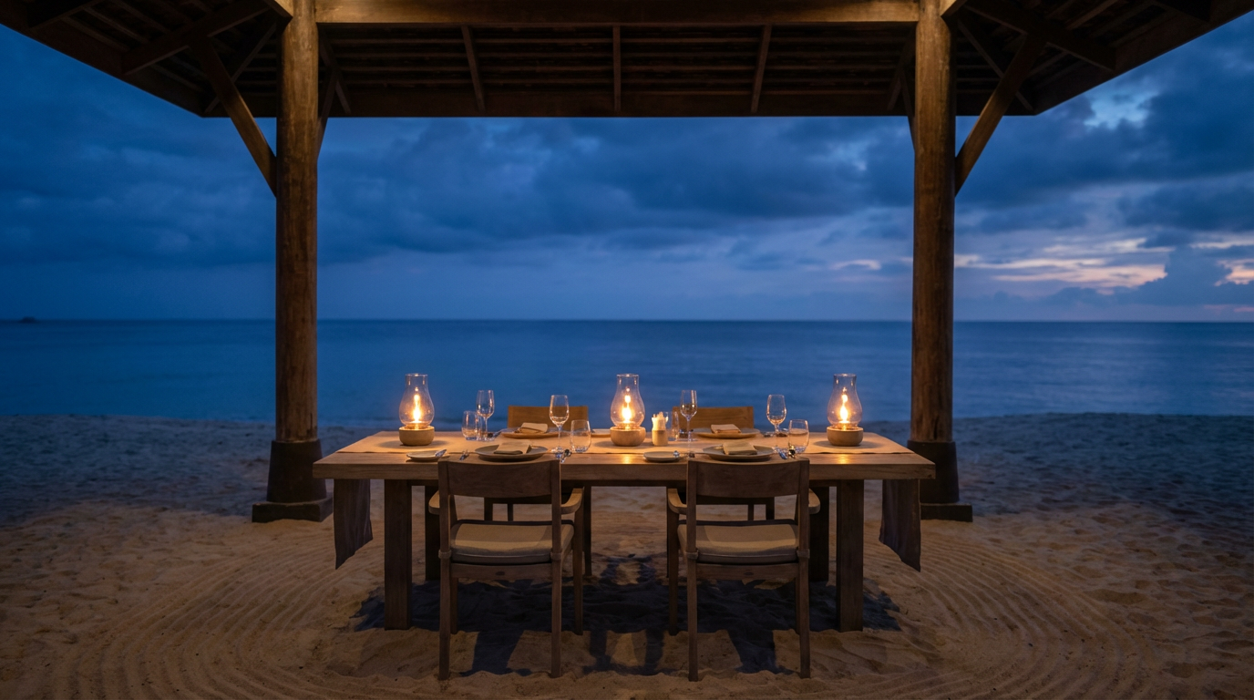 Open-front Layan beach restaurant at dusk with a single long table set for four on raked sand and hurricane lamps