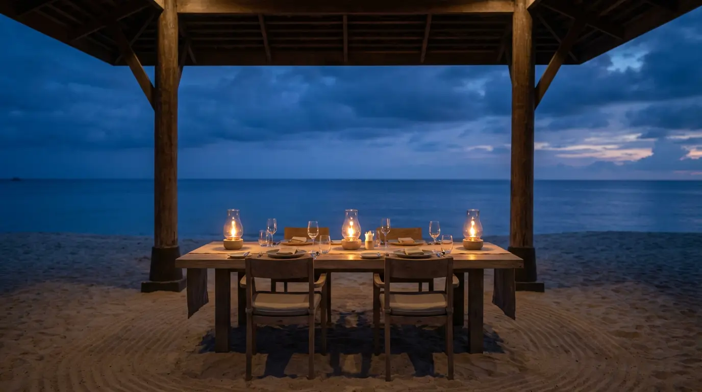 Open-front Layan beach restaurant at dusk with a single long table set for four on raked sand and hurricane lamps
