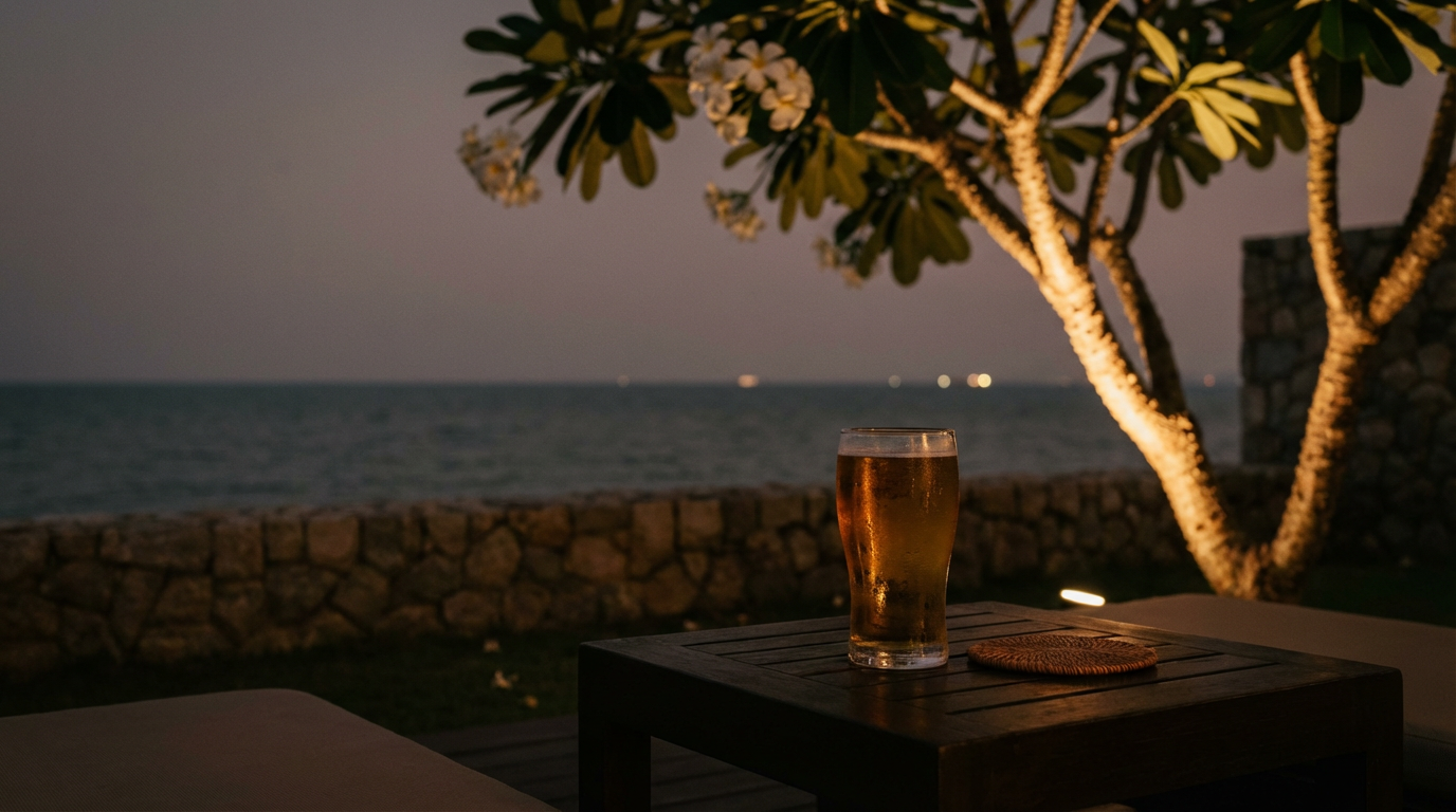 Quiet Hua Hin hotel terrace at dusk with a single cold beer on a low table and the dark gulf beyond a stone wall