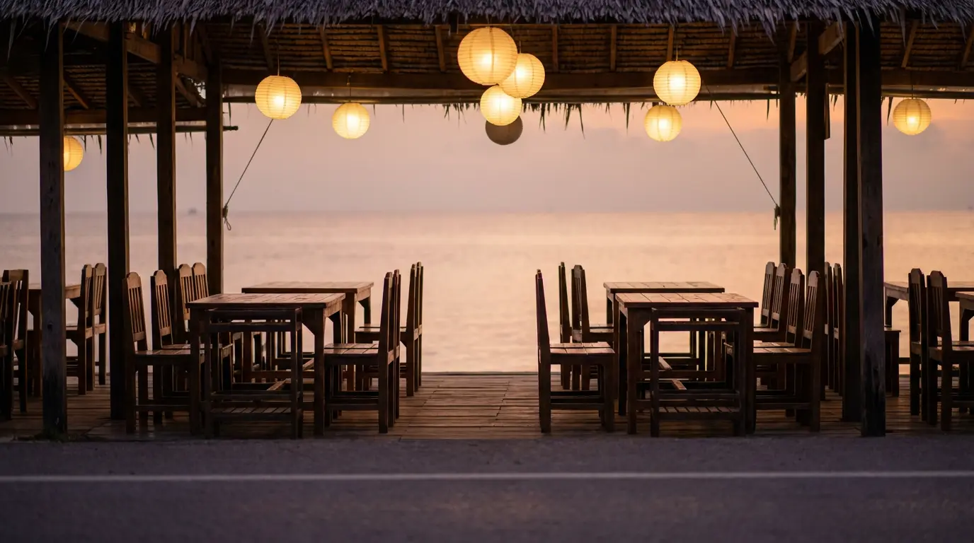 Open-air seafood restaurant at Cha-Am at dusk with paper lanterns just lit and empty wooden tables facing the gulf