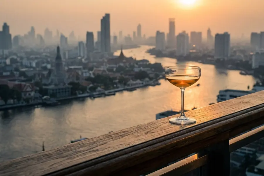 Bangkok rooftop view at dusk with a long temple spire silhouetted against the warm sky and the river curving below