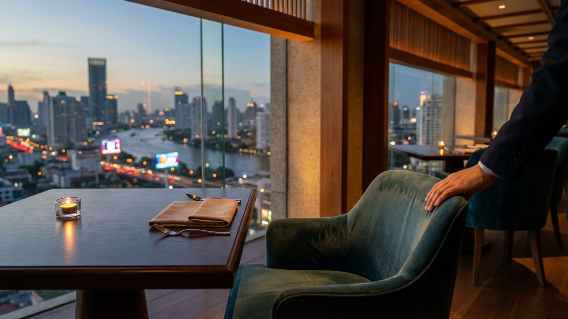 Window-side table at golden hour with the Bangkok skyline beyond, a maitre d' pulling out the velvet chair