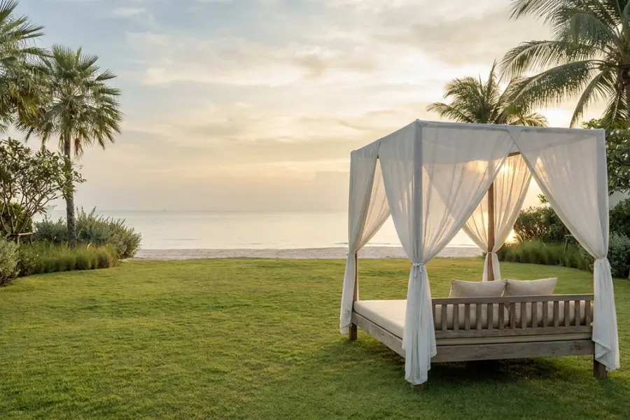 A Hua Hin private villa lawn at dawn with a long teak meeting table set for eight, the Gulf of Thailand softly visible beyond