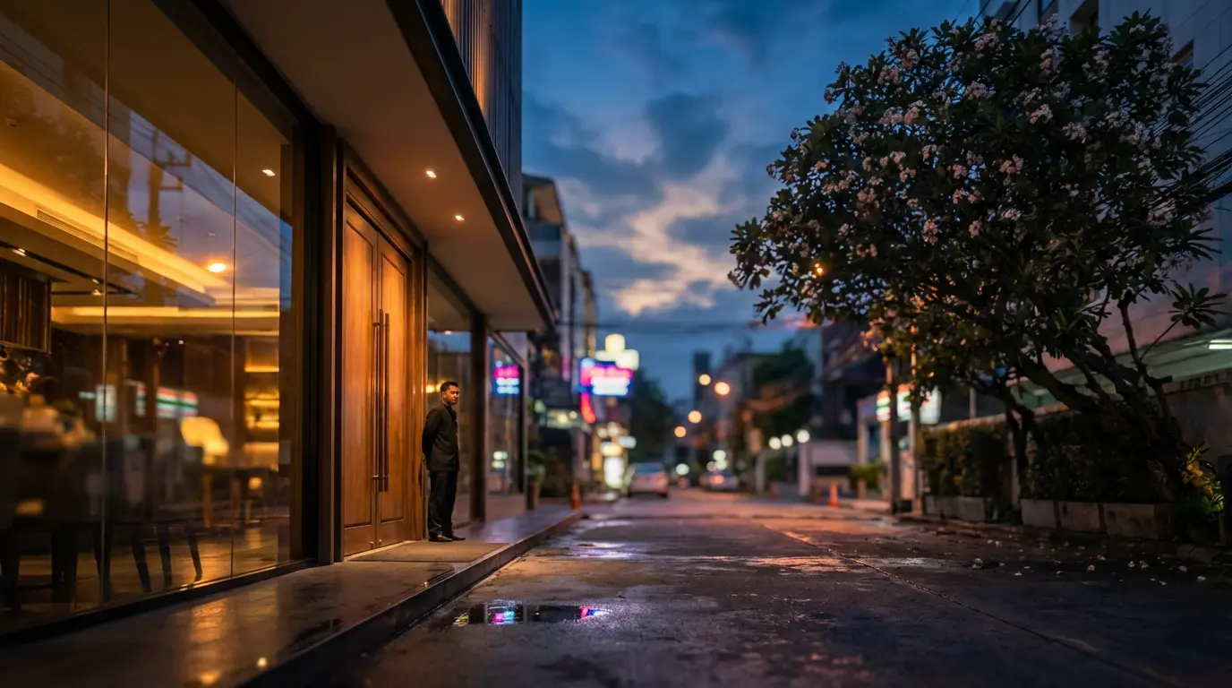 Thonglor side soi at dusk with a polished glass restaurant front, a flowering tree and a single doorman