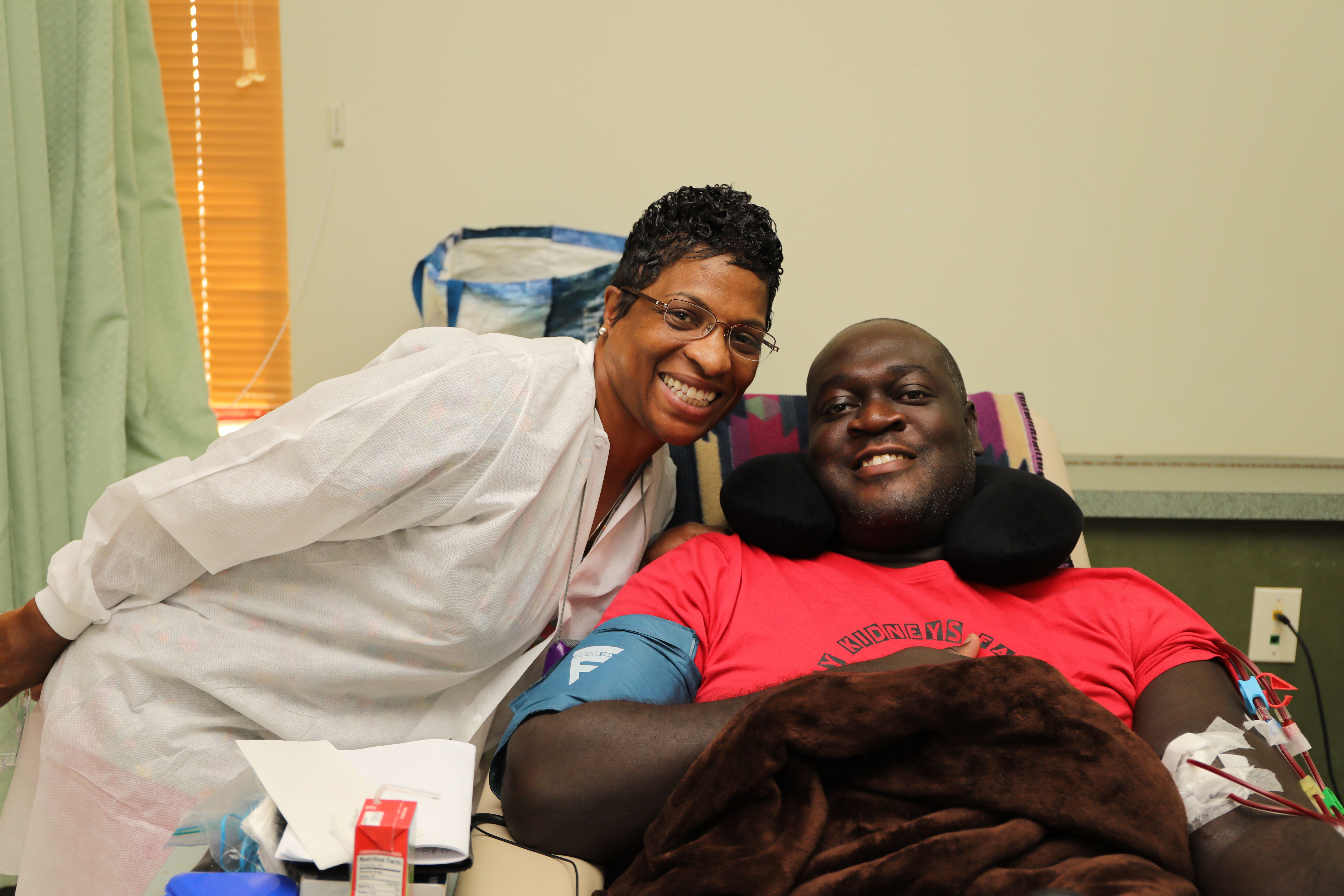 A black female healthcare worker leans in, smiling, to take a photo with a black male patient who is seated.