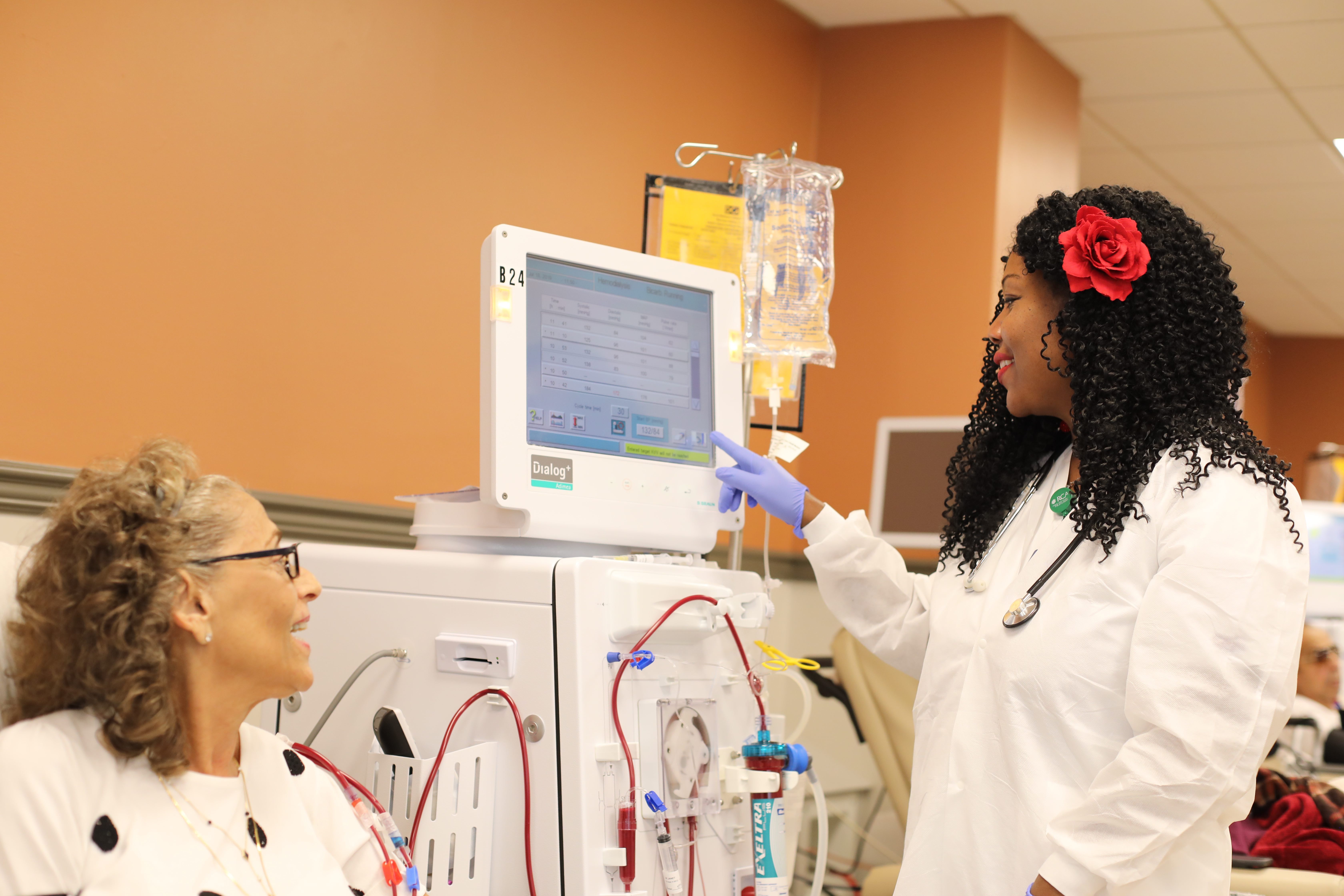 A black female healthcare worker with a red flower in her hair helps a patient at a dialysis station.