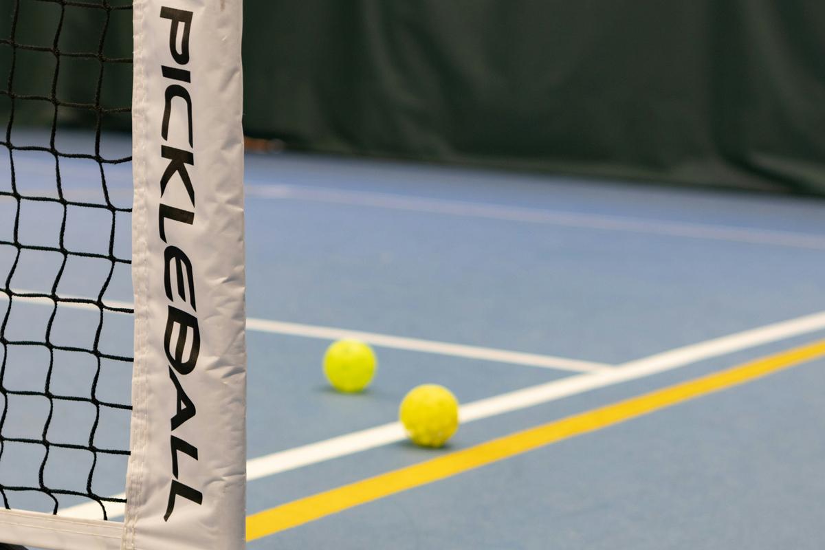 Two yellow pickleballs resting on a blue indoor court near a pickleball net with 'PICKLEBALL' text on the white post, showing the court lines and equipment setup for pickleball game play.
